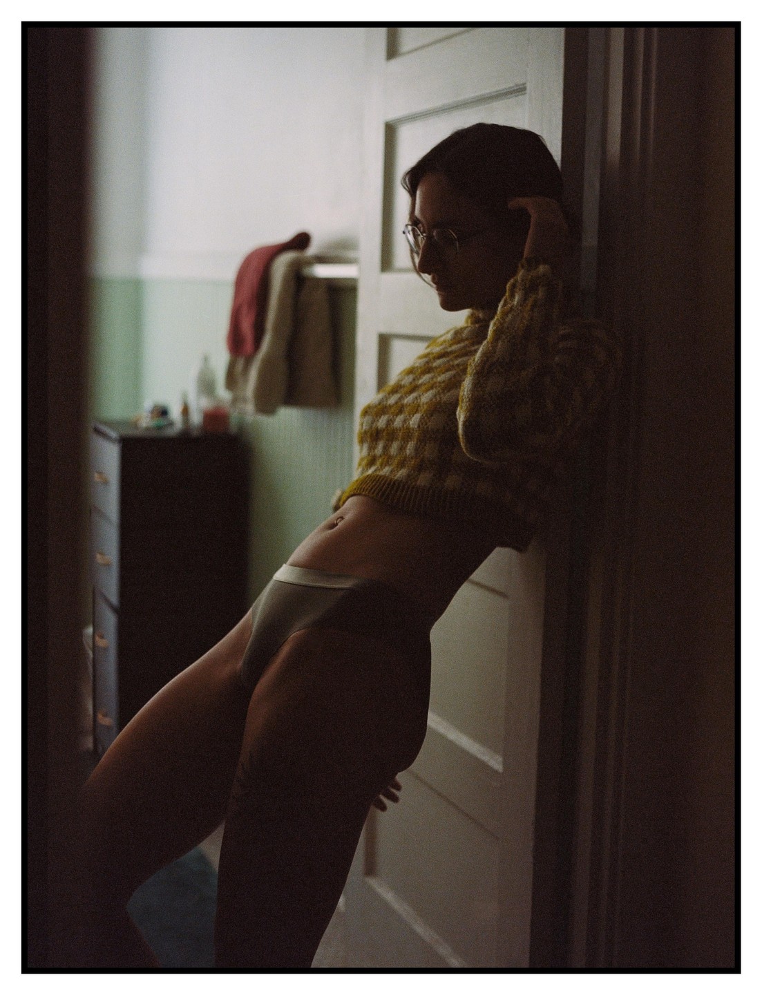 A woman in a sweater and underwear leans against a doorframe in a dimly lit room.
