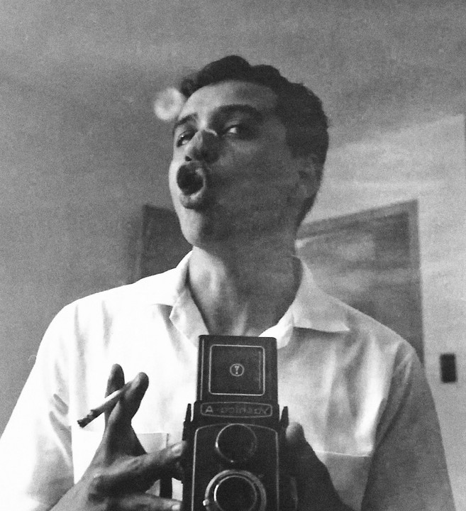 A man with a cigarette in his hand holds a twin-lens reflex camera up to his face, making an O-shape with his mouth as if blowing a smoke ring.