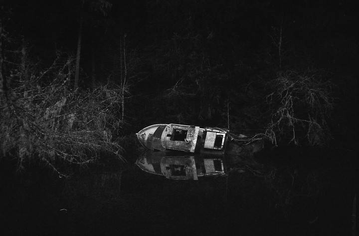 A derelict boat is partially submerged in dark water, reflecting the surrounding trees and the boat itself.
