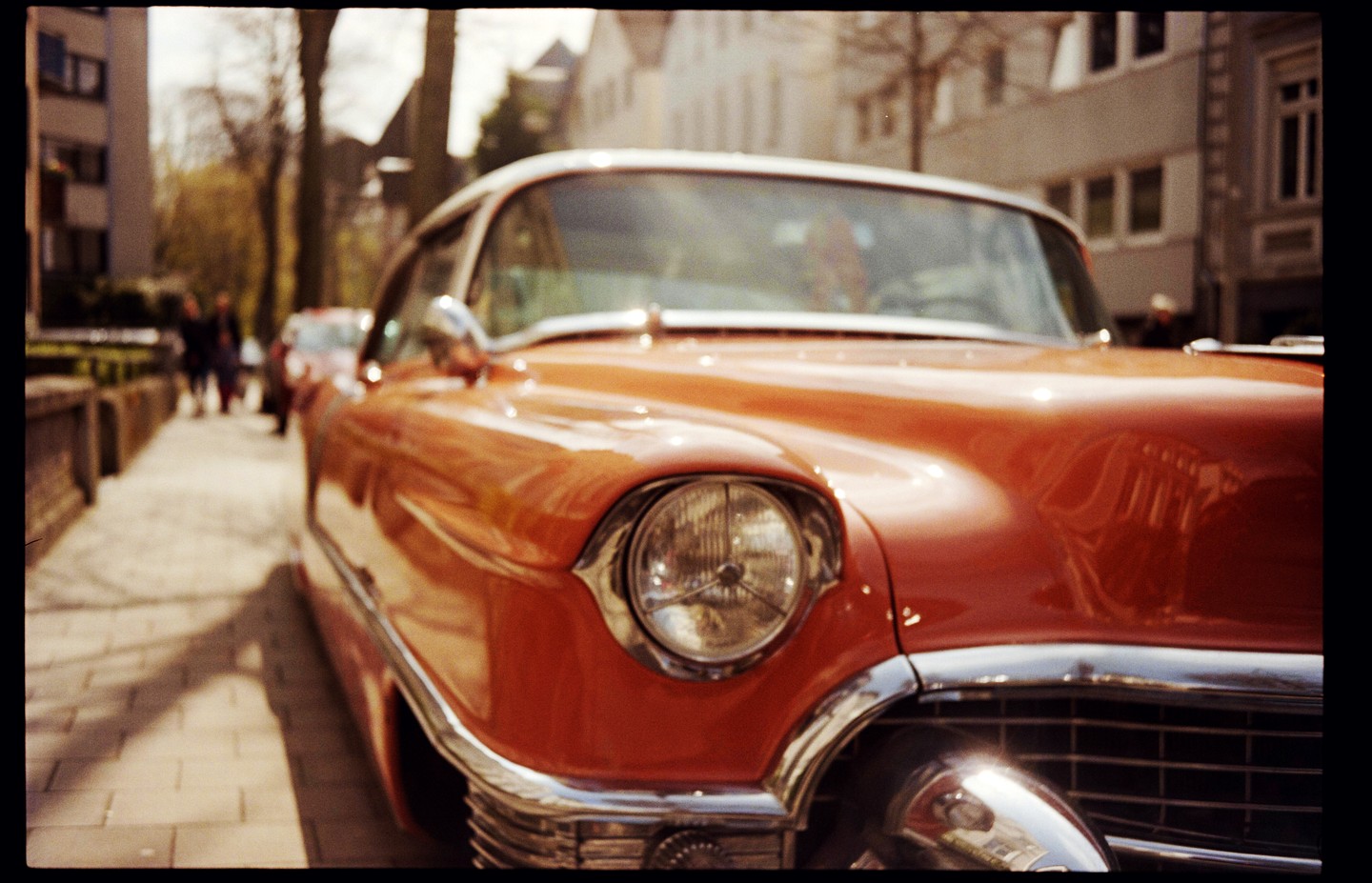 A classic orange and white car is parked on a street with buildings and trees in the background.