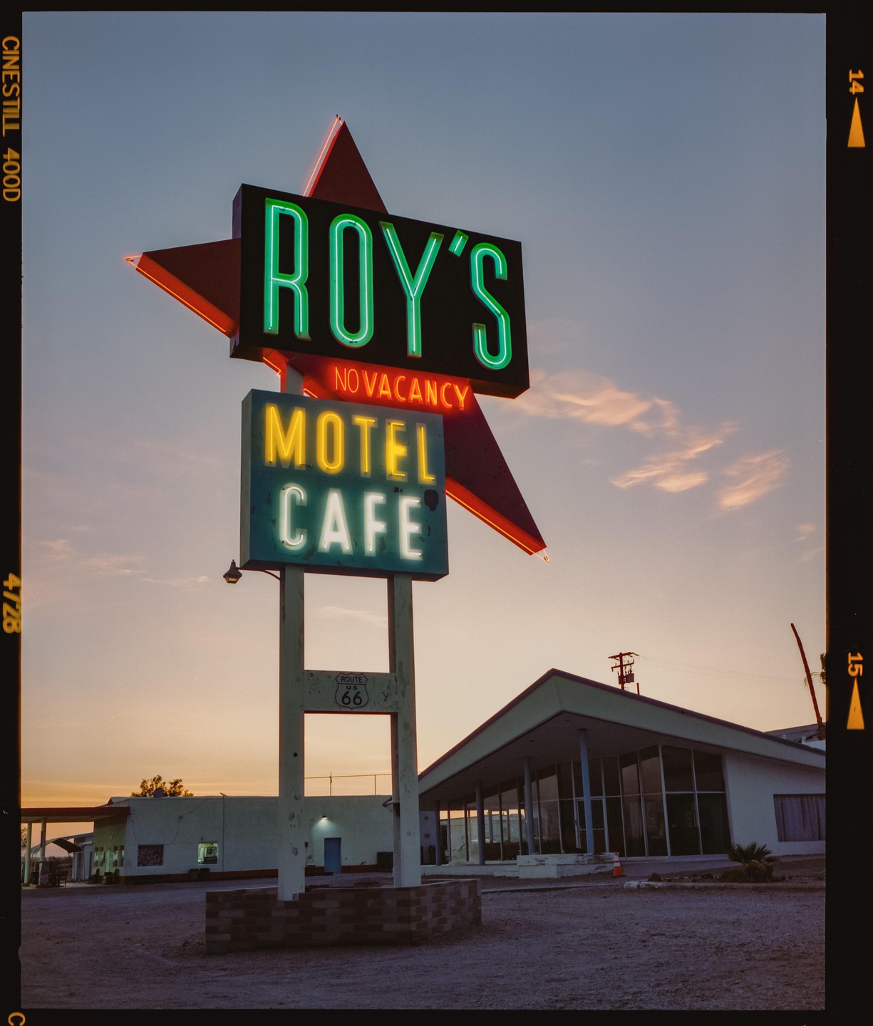 A neon sign for Roy's Motel and Cafe stands against a sunset sky next to a building with large windows.