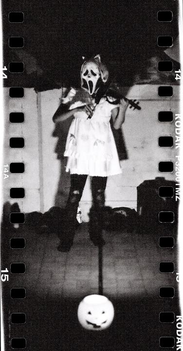 A person in a ghostface mask and white dress plays a violin while standing over a pumpkin pail.