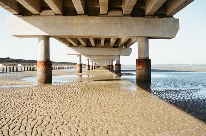 A long bridge extends over a sandy, rippled beach with shallow water reflecting the sky.