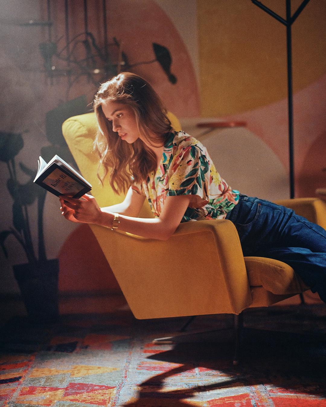 A woman in a floral shirt and jeans is reading a book while relaxing in a yellow armchair.