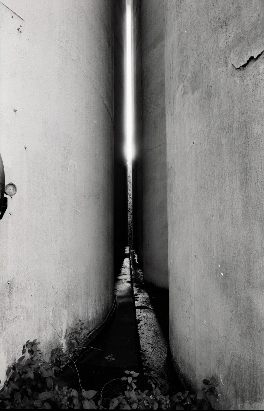 A narrow alleyway between two tall buildings leads to a bright light at the end.
