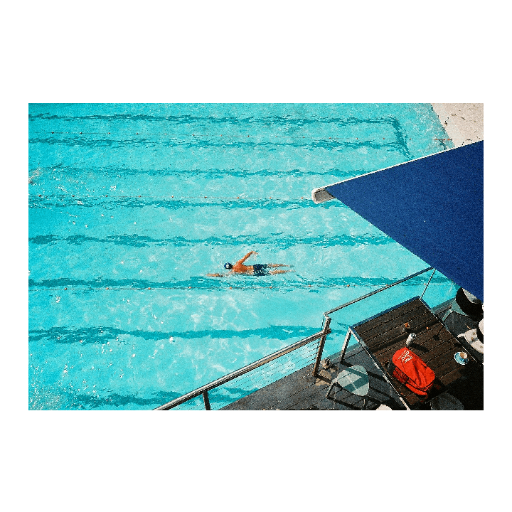 A man swims in a bright blue pool next to a deck with a blue awning.