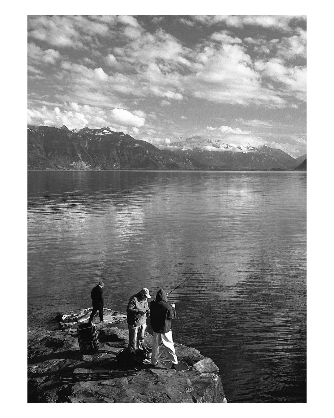 Three men are fishing from a rocky shore with a large lake and mountains in the background under a cloudy sky.