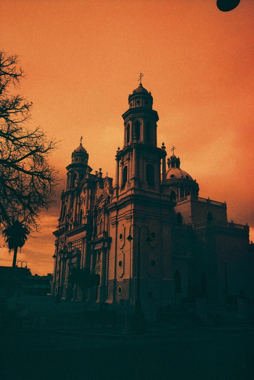 A large, ornate church with multiple towers and domes stands silhouetted against an orange sky.