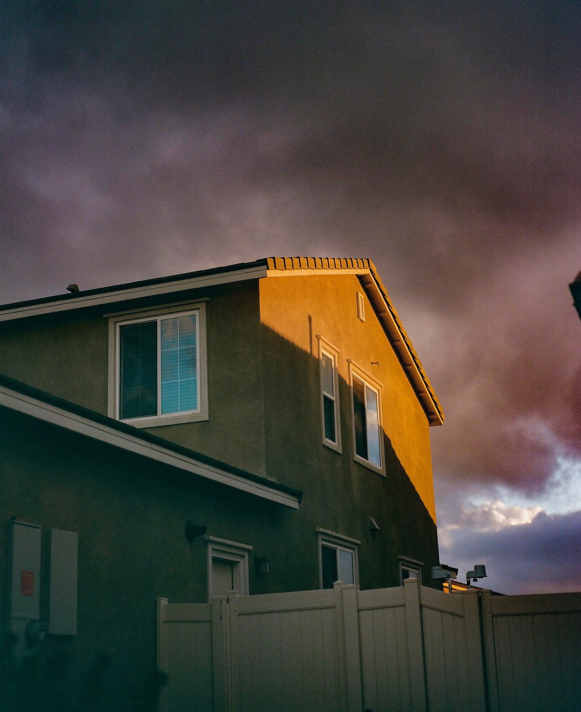 A house with a fence in front of it is illuminated by the setting sun under a cloudy sky.