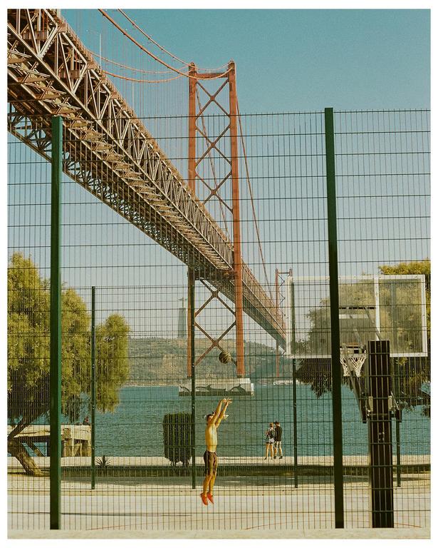 A man jumps to shoot a basketball on an outdoor court next to a body of water with a large bridge overhead.
