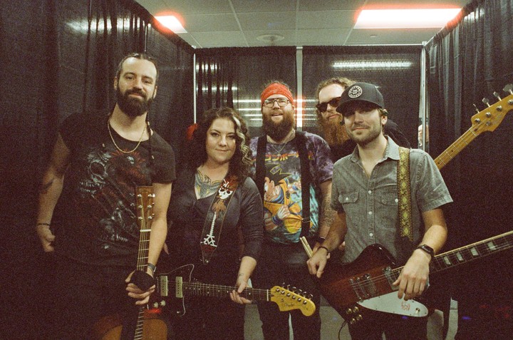 Five musicians, three men and two women, pose with their instruments in a backstage area.