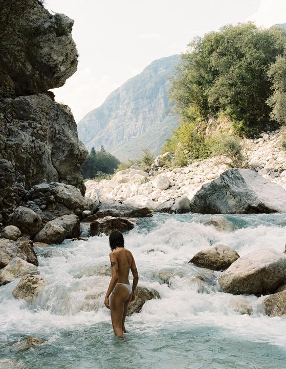 A woman stands in a rocky river with mountains and trees in the background.