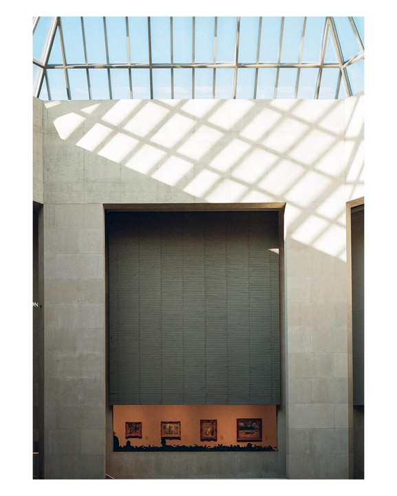 Sunlight streams through a glass ceiling onto a museum wall above a display of paintings.