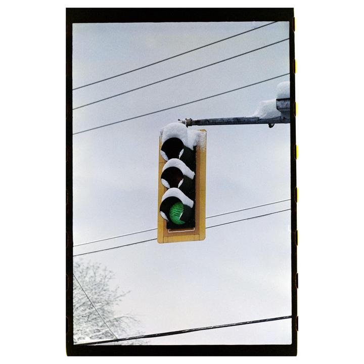 A traffic light covered in snow hangs against a cloudy sky with power lines and a snow-dusted tree visible.