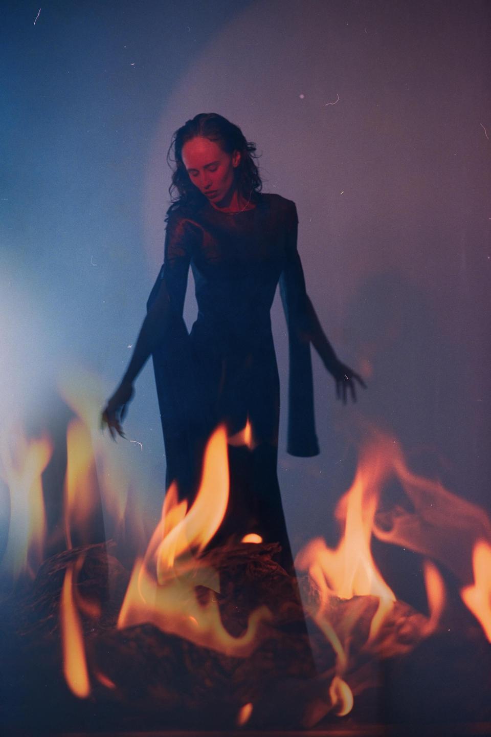 A woman in a long dress stands with flames in the foreground.