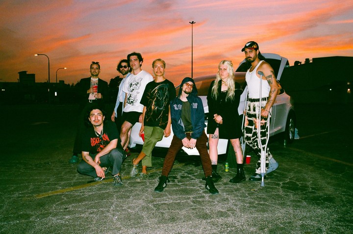 A group of eight people pose in a parking lot at sunset with a white car behind them.