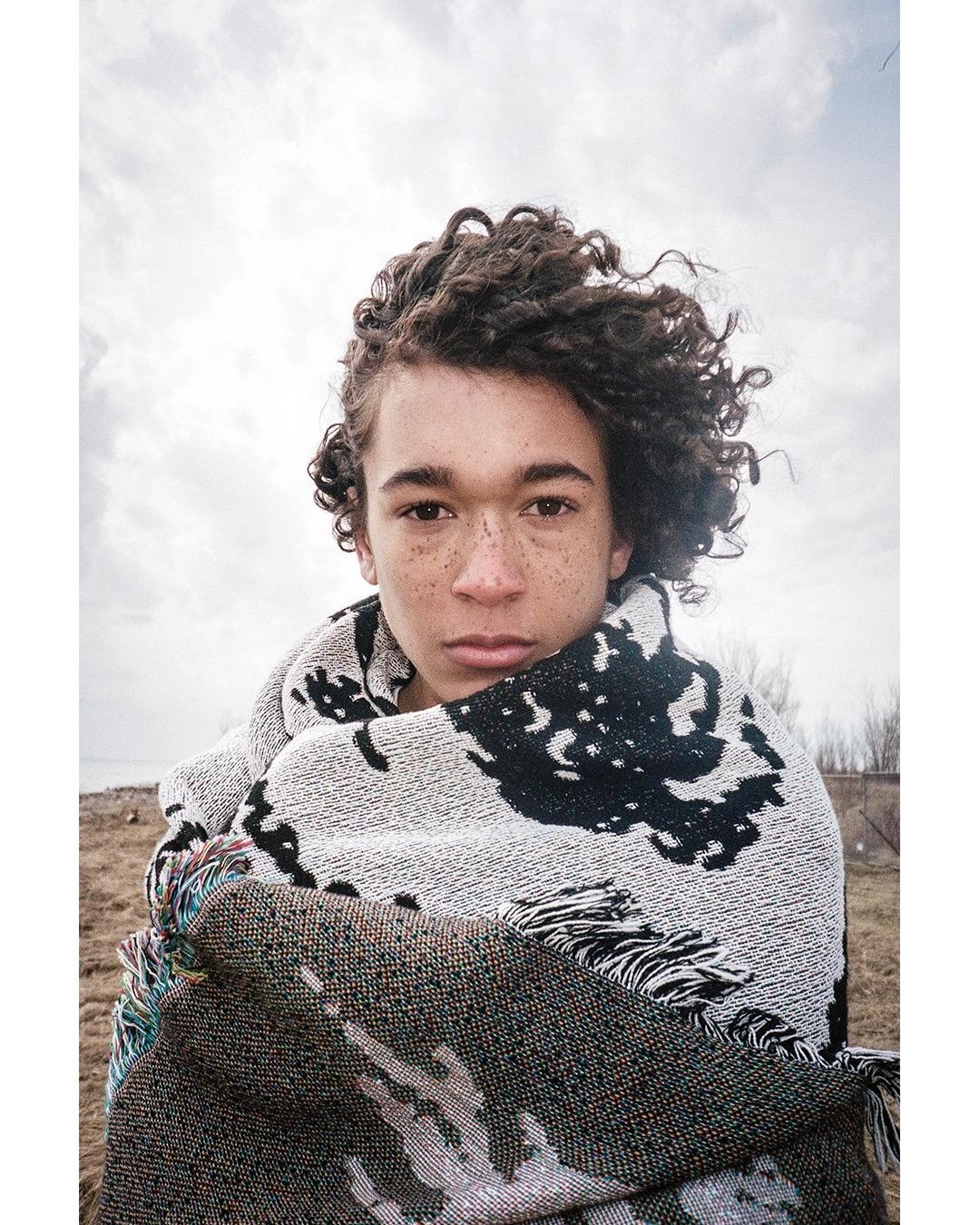 A boy with curly hair and freckles is wrapped in a patterned blanket.