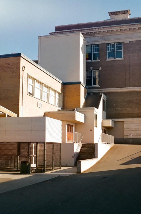 A school building with multiple architectural styles and levels is illuminated by sunlight, casting shadows on its facades and the ground.