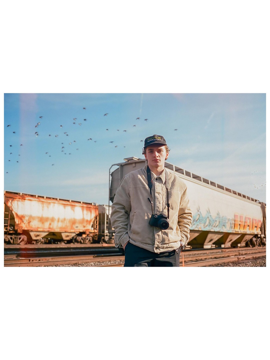 A man stands on train tracks in front of two train cars with birds flying in the sky.