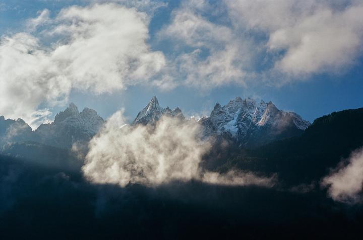 Clouds partially obscure a range of snow-capped mountains under a blue sky.