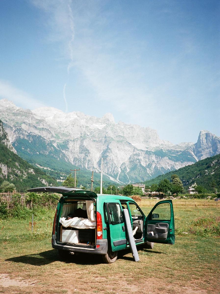 A green van with its doors open is parked in a grassy field with mountains in the background.