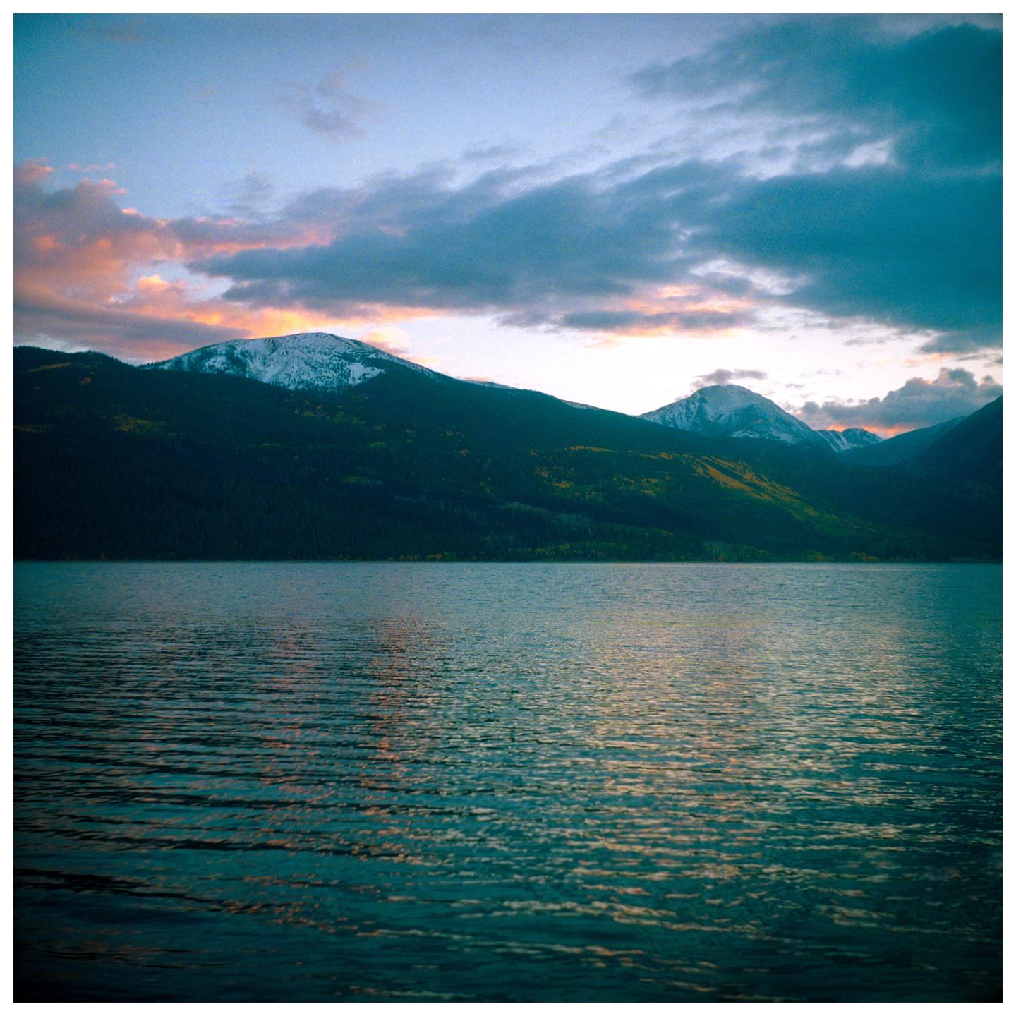 A body of water reflects the sky and mountains with snow-capped peaks under a cloudy sky at dusk.