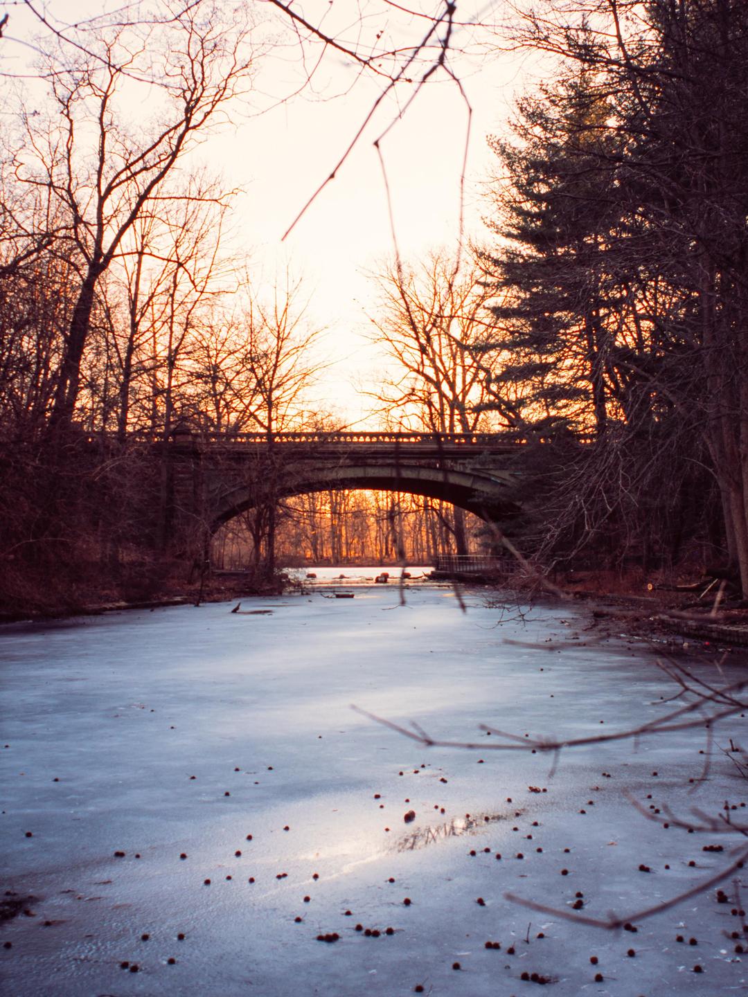 A frozen river flows under a stone bridge surrounded by bare trees at sunset.