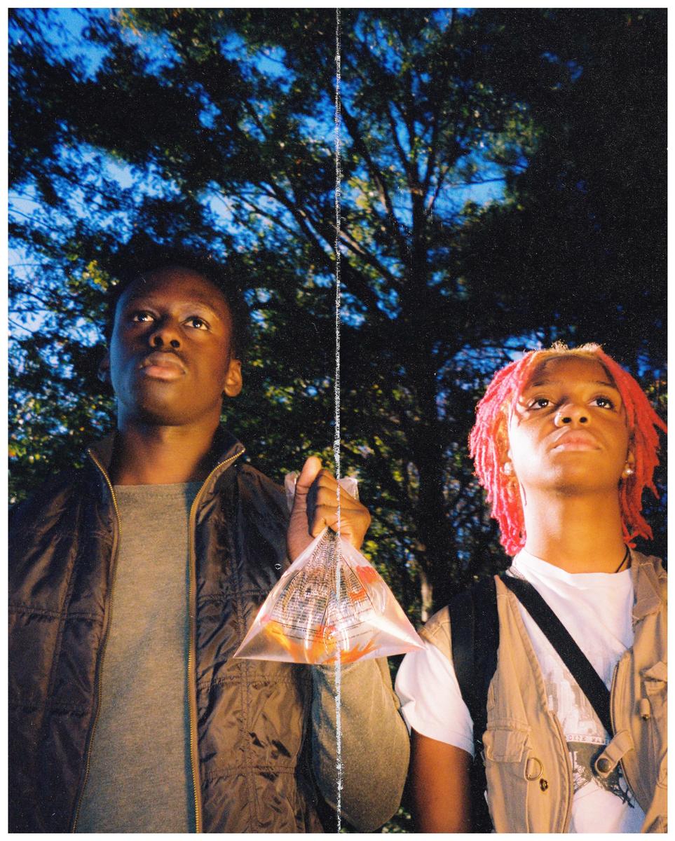 Two young men stand outdoors at night, one holding a plastic bag with goldfish.