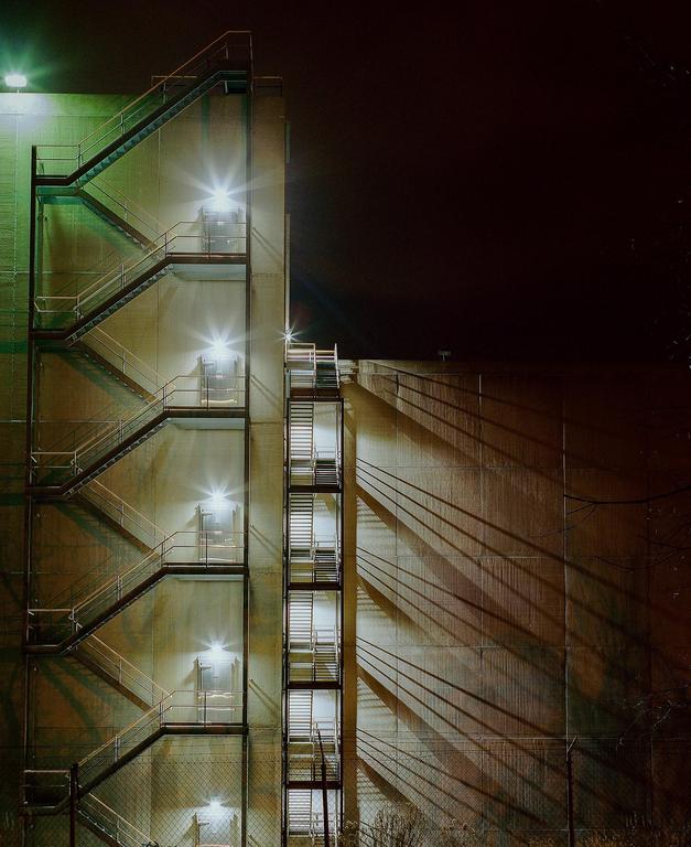 Two fire escapes with bright lights cast long shadows on the side of a building at night.