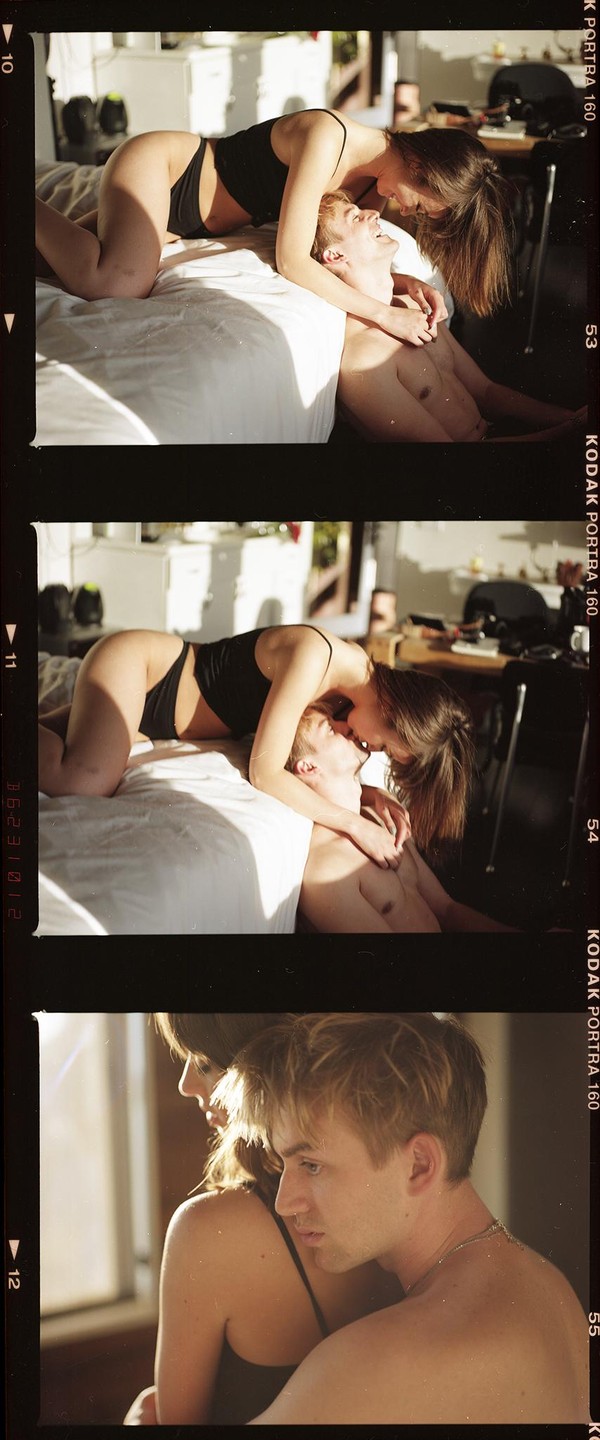 A woman in a black top and underwear is on top of a man lying on a bed, with sunlight streaming in.