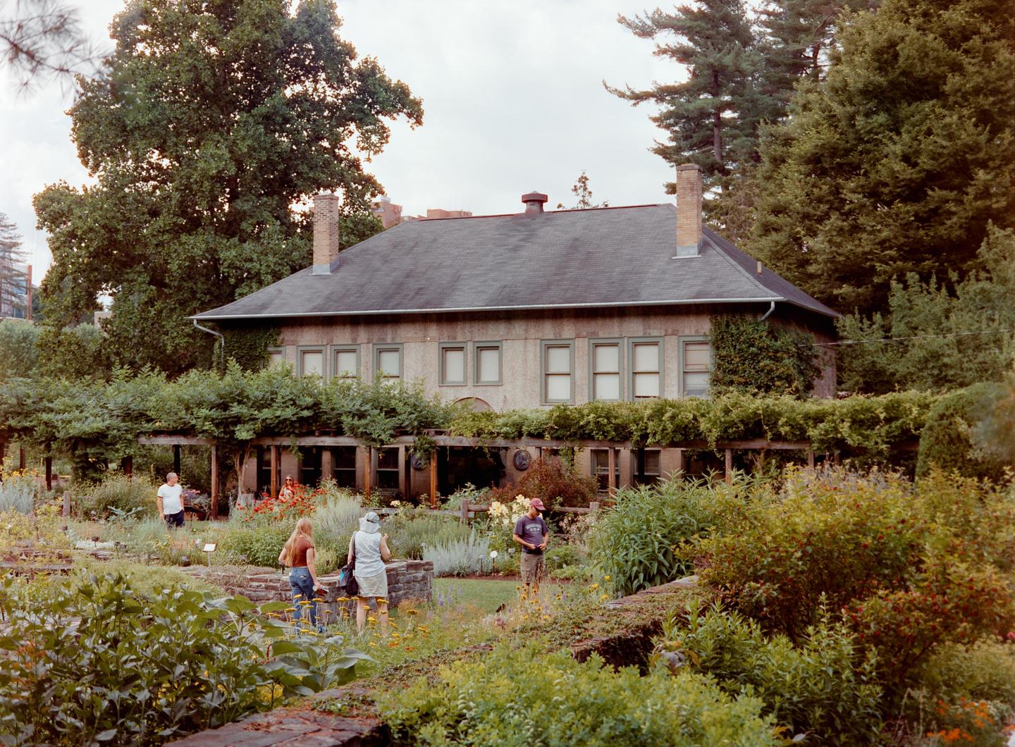 A large house with a dark roof and many windows is surrounded by lush green gardens and trees, with several people walking through the garden.