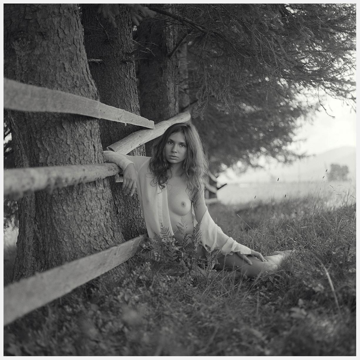 A woman with long hair sits in tall grass next to a wooden fence and a tree.