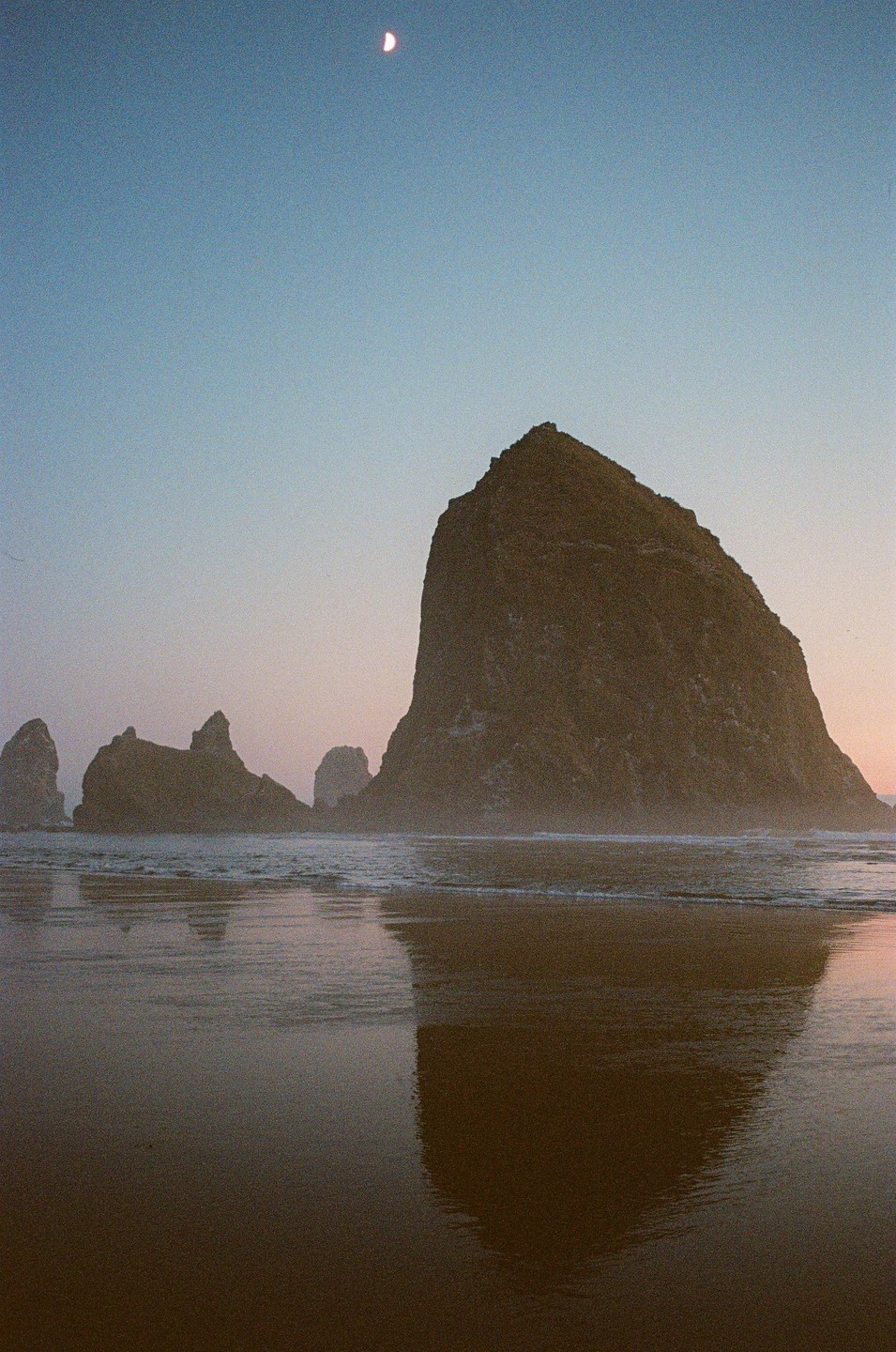 A crescent moon hangs in the sky above a large rock formation on a beach with its reflection visible in the wet sand.