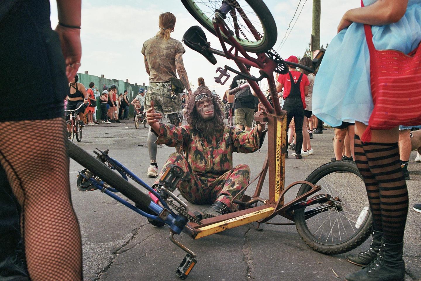 A person in a gorilla mask and camouflage jumpsuit sits on the ground next to a bicycle, surrounded by other people.