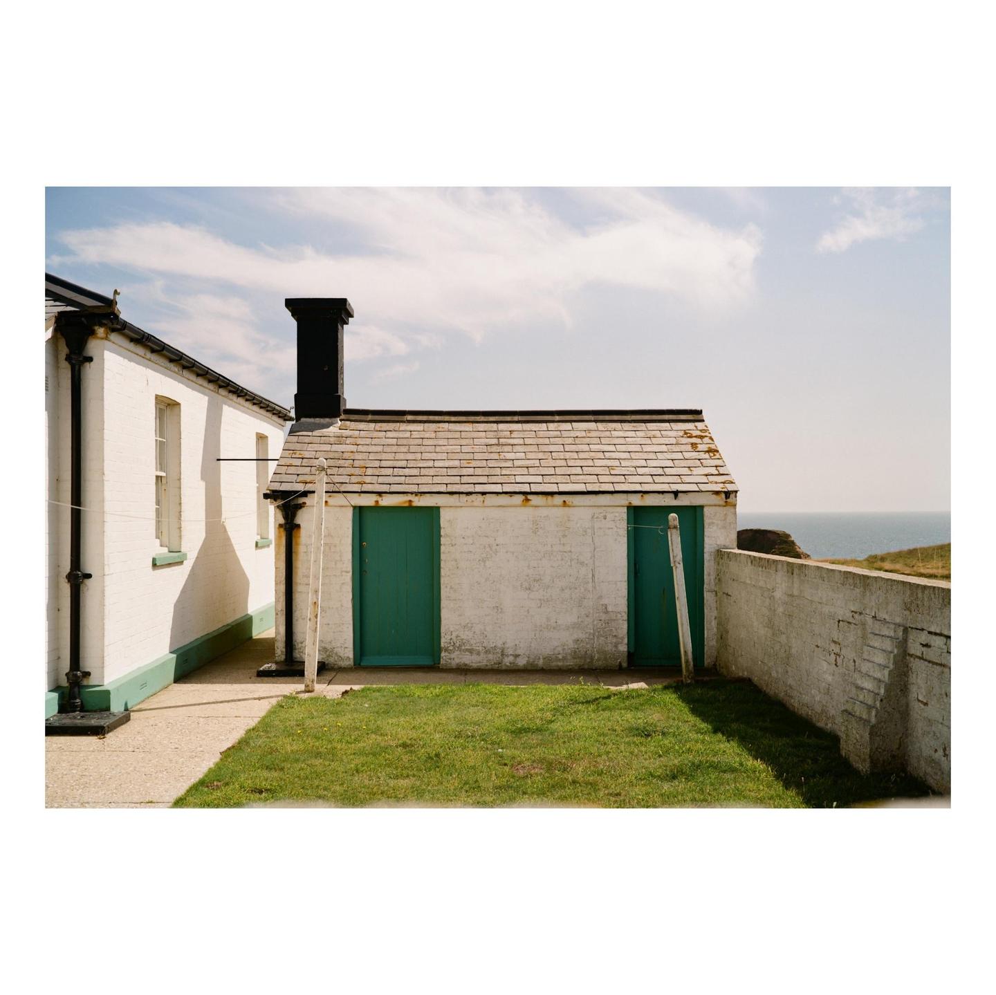 A small white building with two green doors and a black chimney stands next to a larger white building, with the ocean visible in the distance.