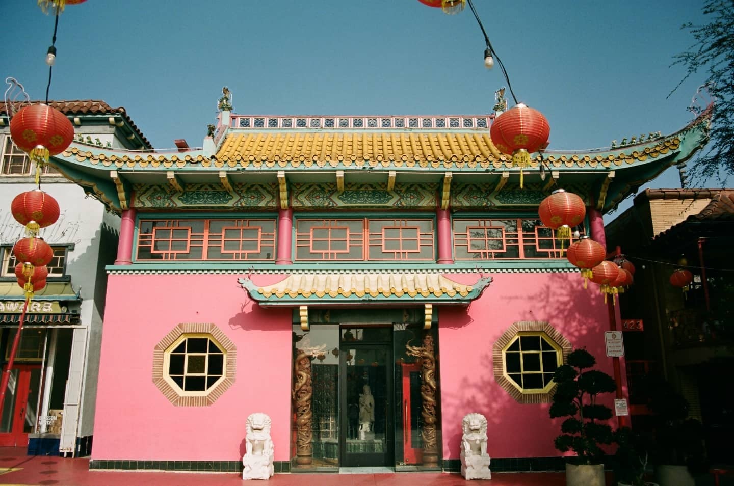 A bright pink building with a yellow and blue tiled roof is adorned with red lanterns under a clear blue sky.