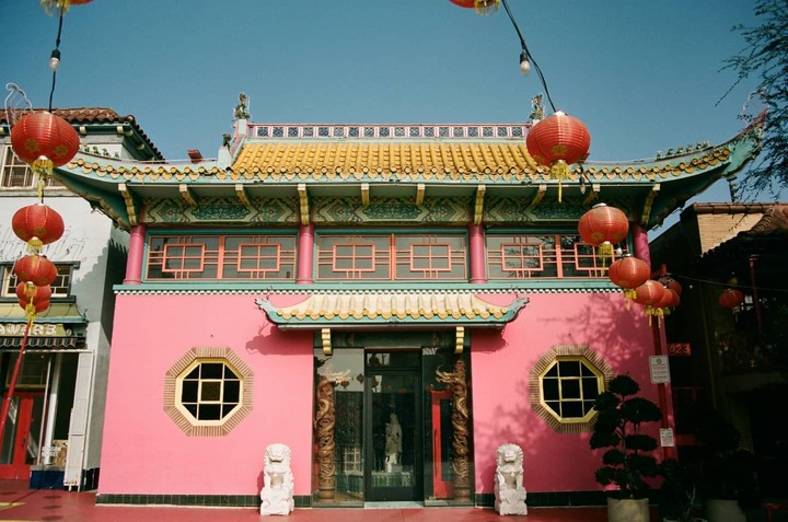 A bright pink building with a yellow and blue tiled roof is adorned with red lanterns under a clear blue sky.
