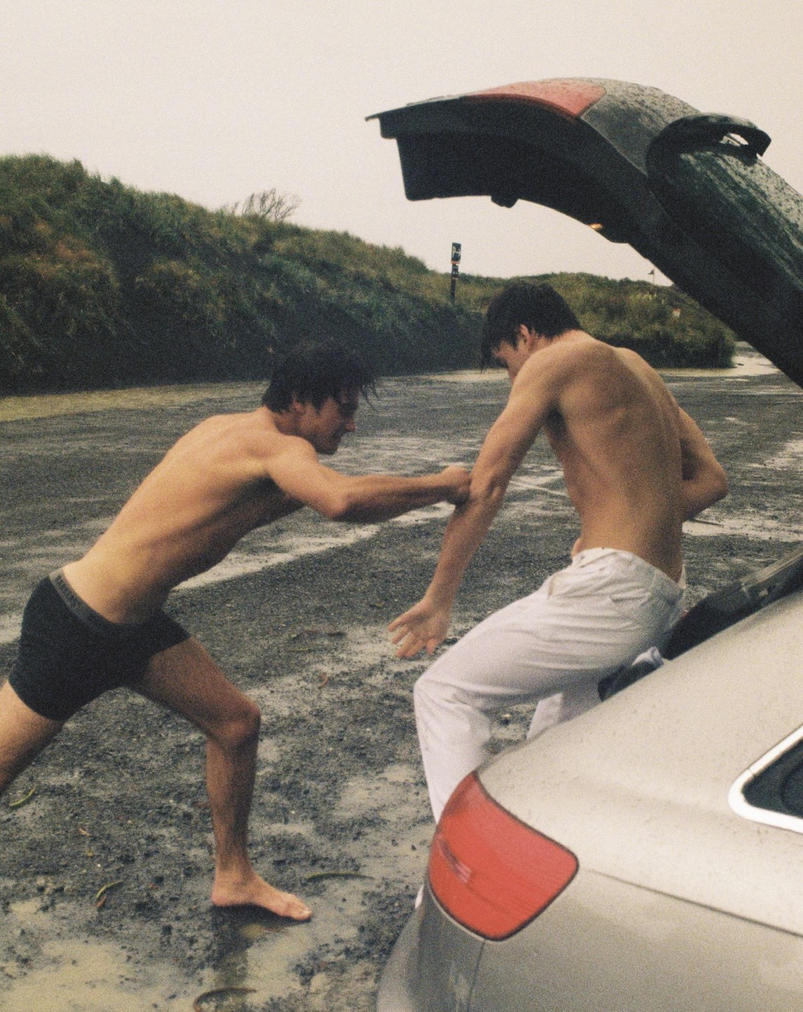 Two shirtless men are standing by an open car trunk on a wet, gravelly surface next to a grassy hill.