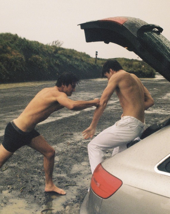 Two shirtless men are standing by an open car trunk on a wet, gravelly surface next to a grassy hill.