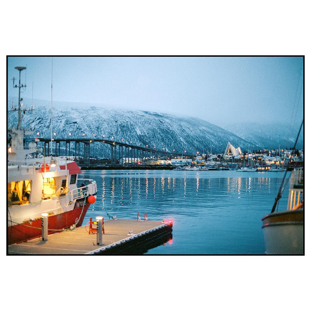 A boat is docked in a harbor with a snowy mountain and a bridge in the background.