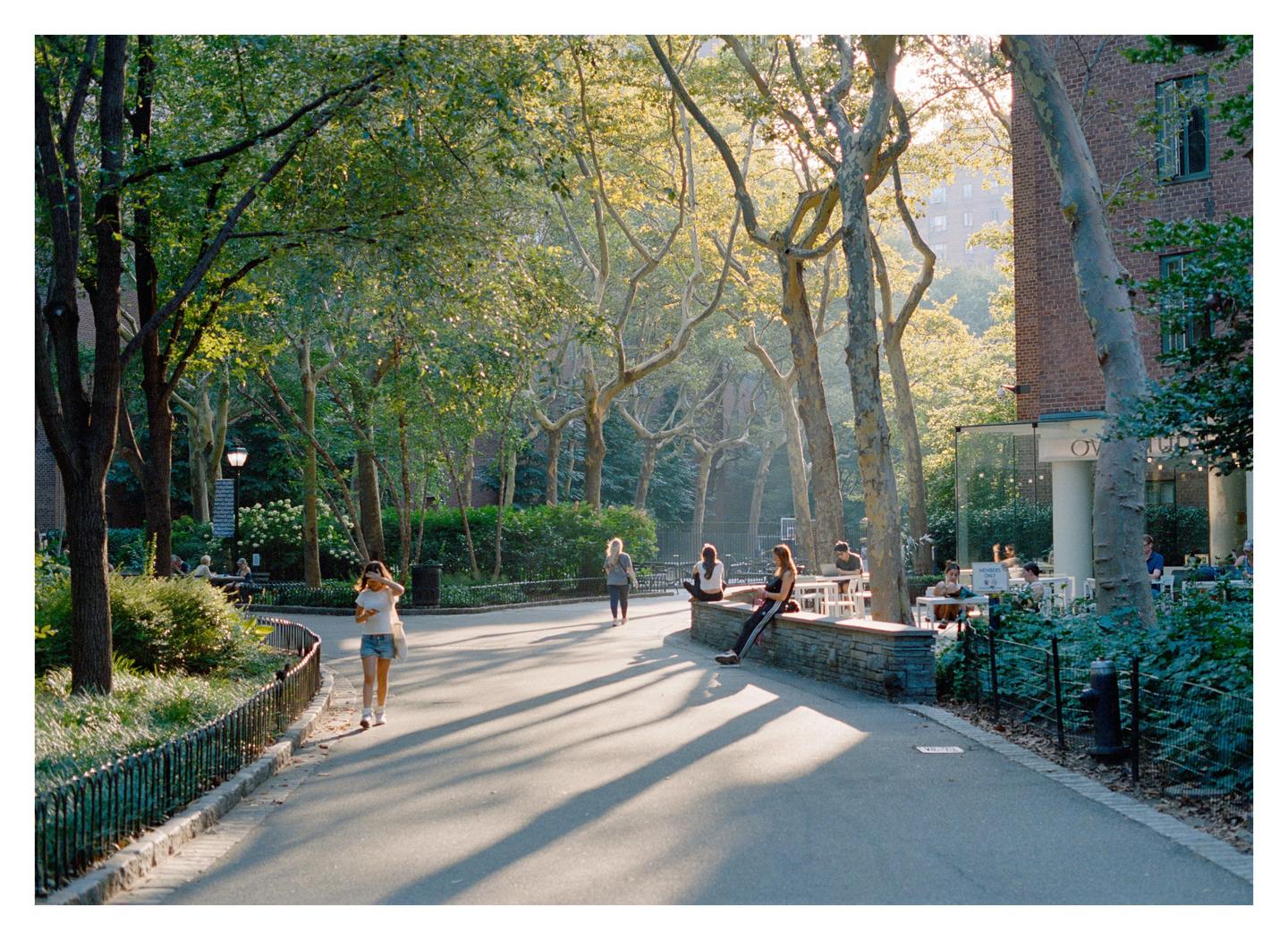 A woman walks along a tree-lined path with long shadows while other people sit at tables in the background.