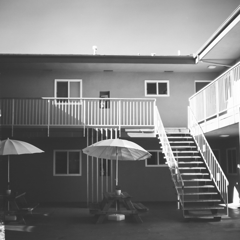 Two patio umbrellas shade picnic tables in the courtyard of a building with external stairs and balconies.