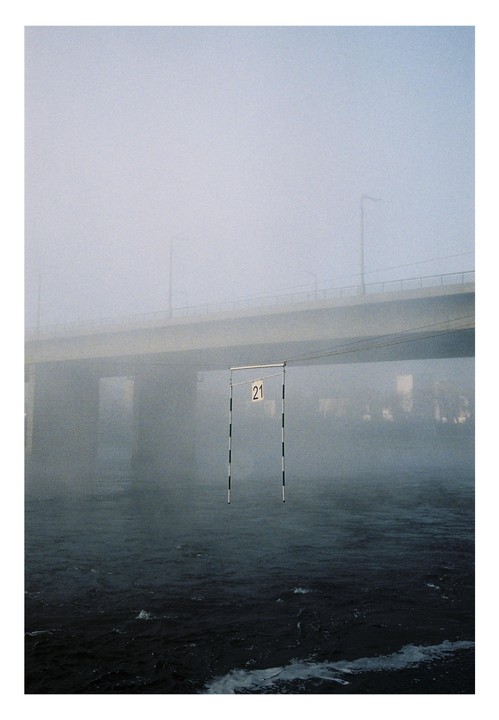 A striped gate with the number 21 hangs over a body of water with a bridge in the background, all shrouded in fog.