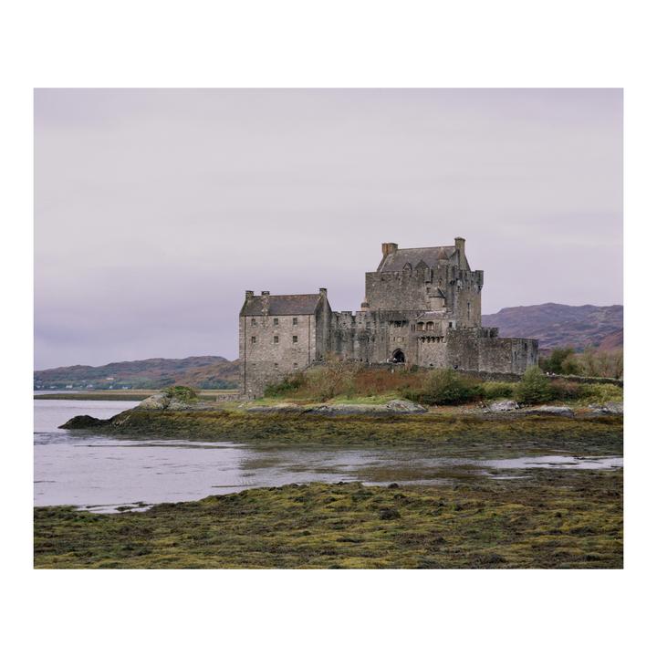 A large stone castle stands on a small island surrounded by water and green land under a cloudy sky.