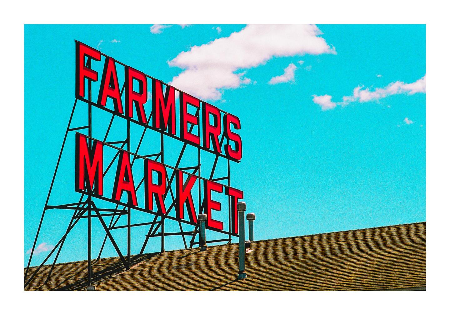 A red "Farmers Market" sign stands against a bright blue sky with a few white clouds, above a brown shingled roof.