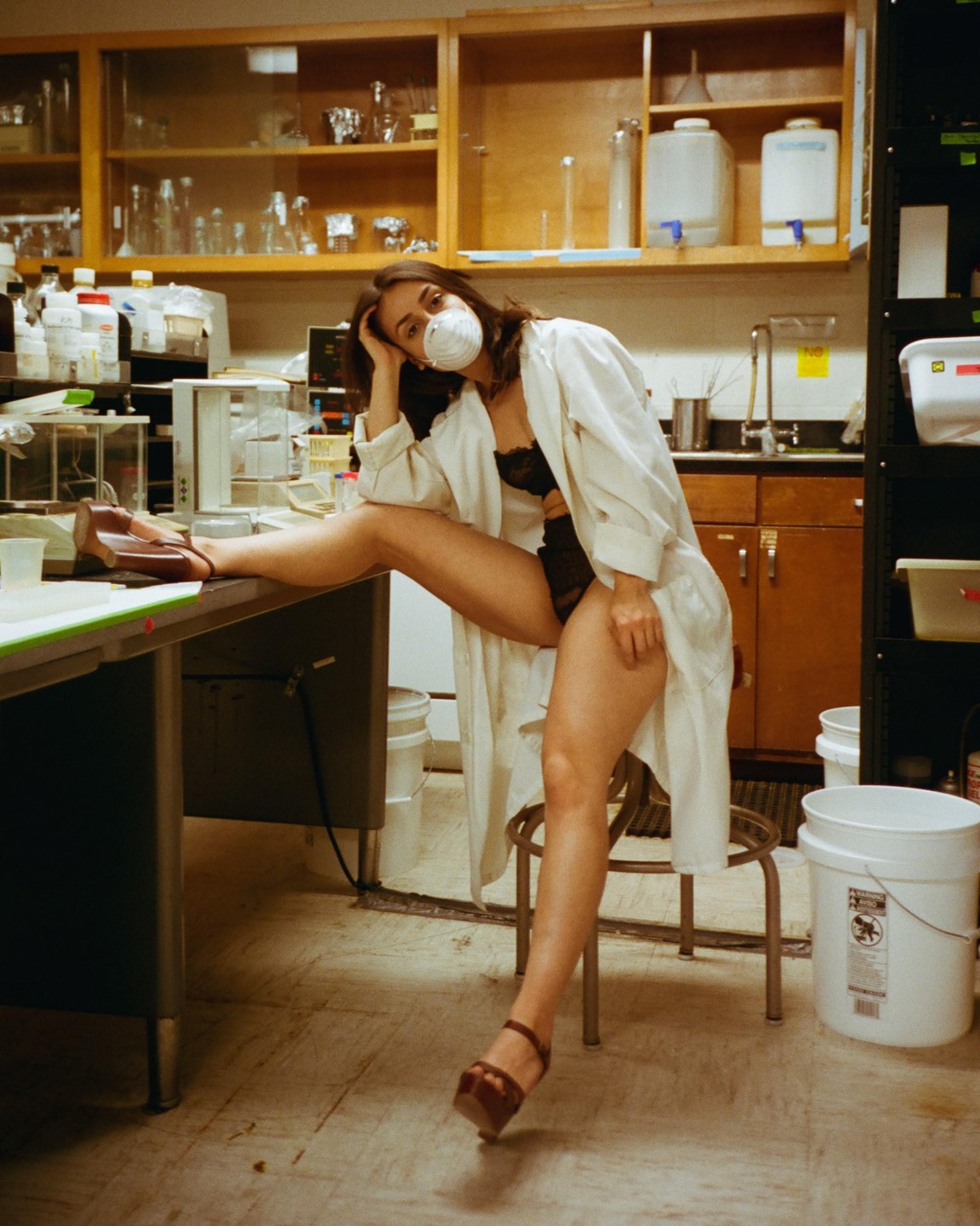 A woman in a lab coat and face mask poses in a laboratory with one leg on a table.