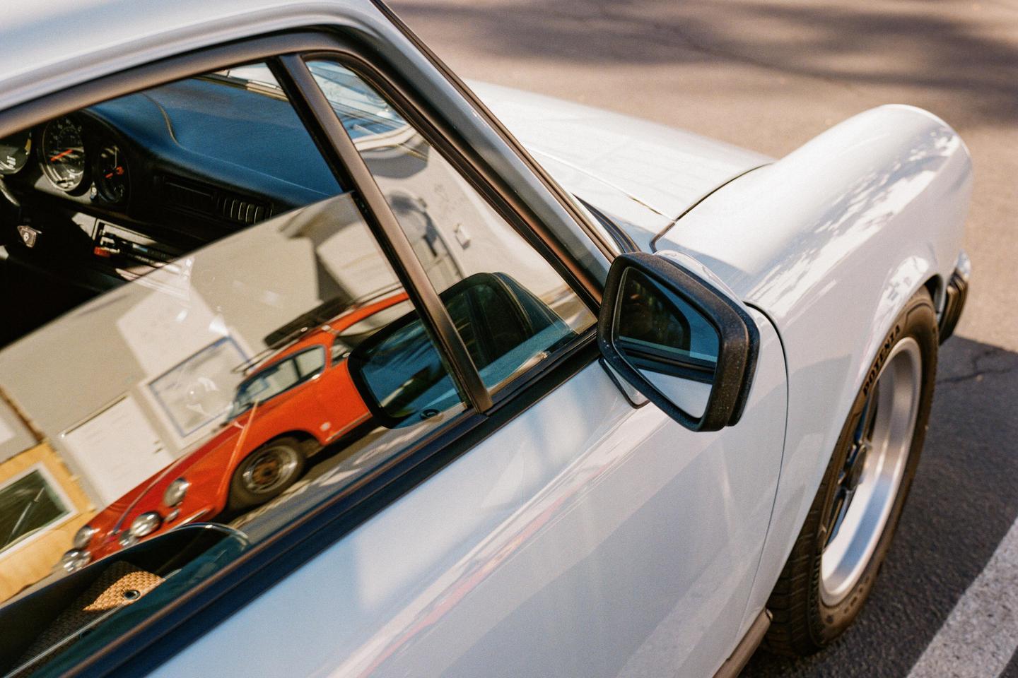 A white car is parked with a reflection of an orange car visible in its window.