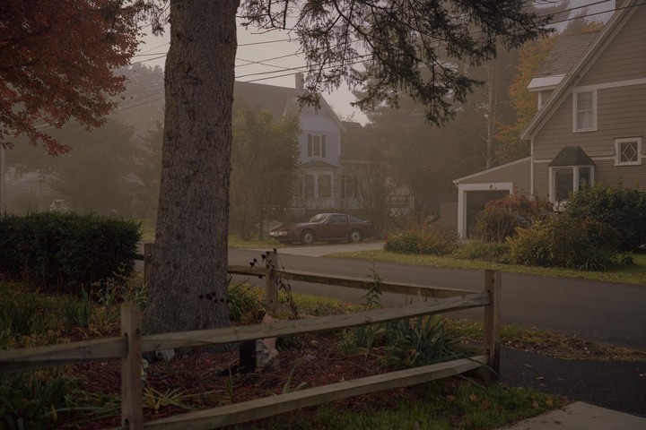 A red car is parked on a street in front of houses on a foggy autumn day.