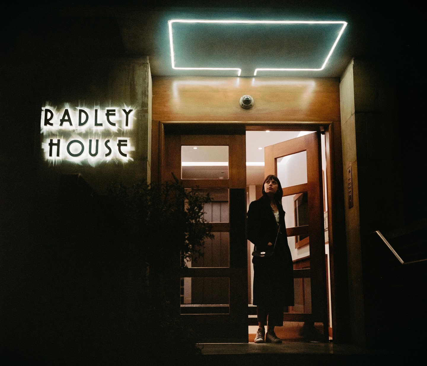 A woman stands in a doorway next to a neon sign that reads "RADLEY HOUSE" at night.