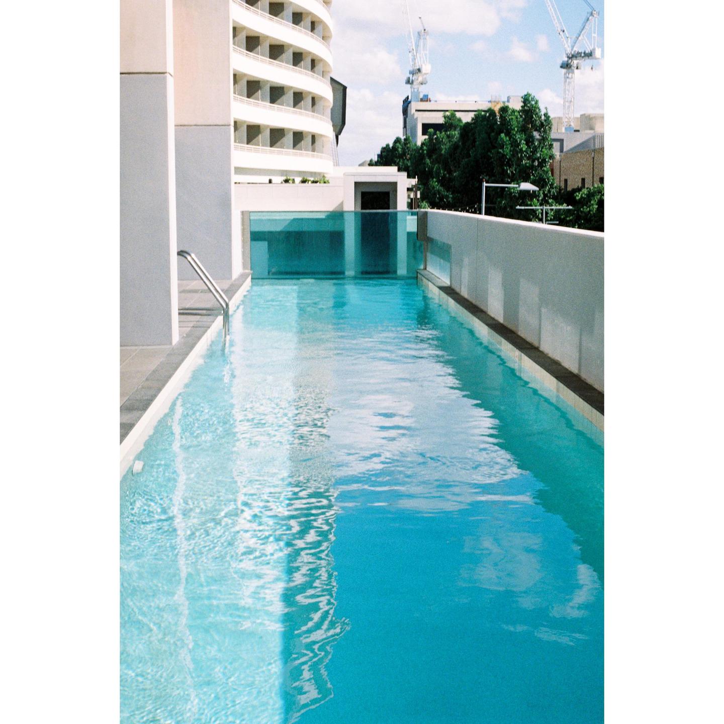 A long, narrow swimming pool is surrounded by modern buildings and trees under a bright sky.